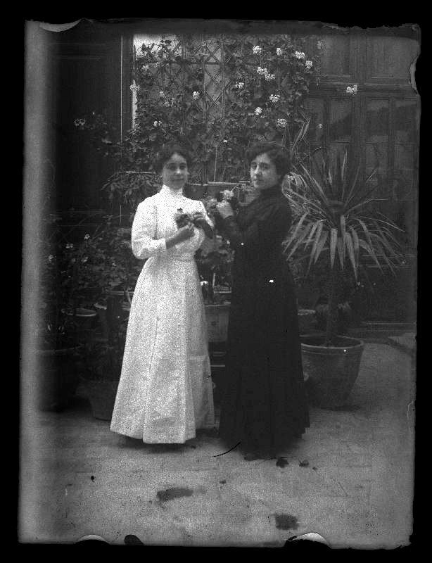 Retrato de cuerpo entero de Angelita Boldrich y Josefa Marín en un patio con flores en las manos.