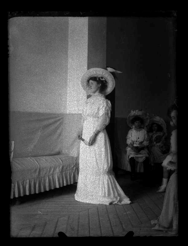 Retrato de cuerpo entero de María Giménez Castellanos vestida de blanco y sombrero en un interior, con varias niñas al fondo