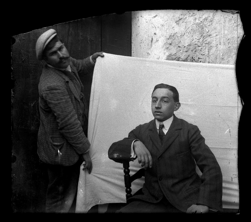 Retrato de Benito López Ruano sentado en una silla mientras un hombre a su lado sujeta una sábana a modo de telón de fondo.