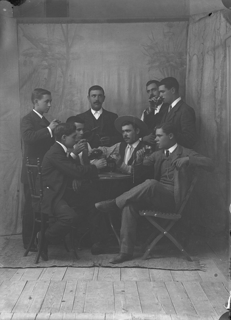 Retrato de estudio de ocho hombres jóvenes bebiendo en torno a una mesa