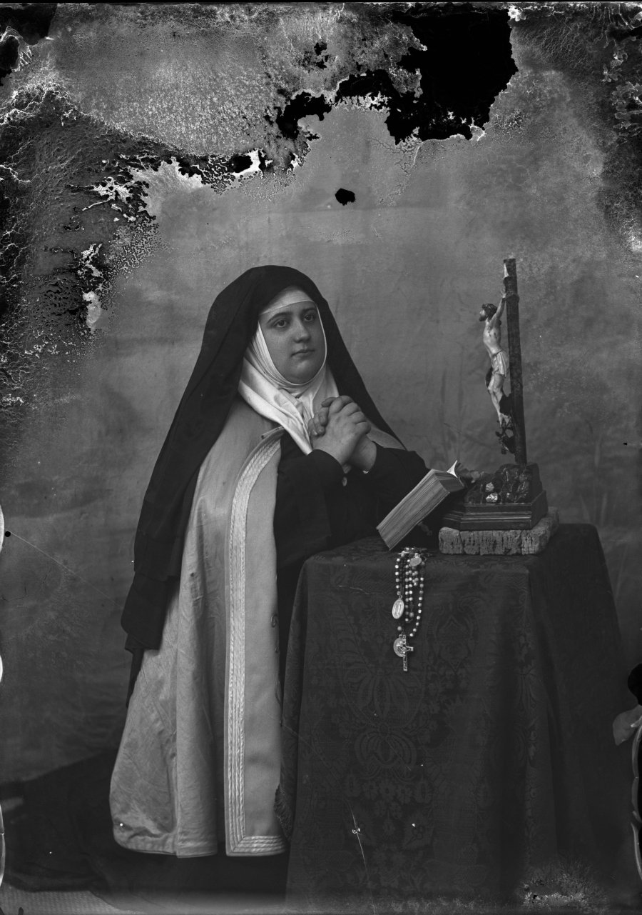 Retrato de estudio de una monja arrodillada en actitud de oración junto a un crucifijo