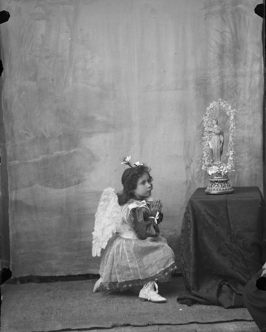 Retrato de estudio de una niña disfrazada de ángel en actitud de oración junto a una figura de la Virgen