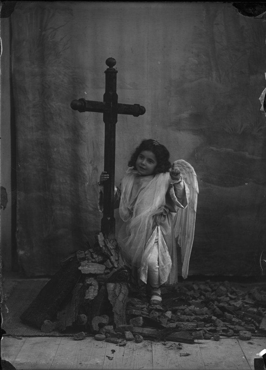 Retrato de estudio de una niña disfrazada de ángel junto a un crucifijo