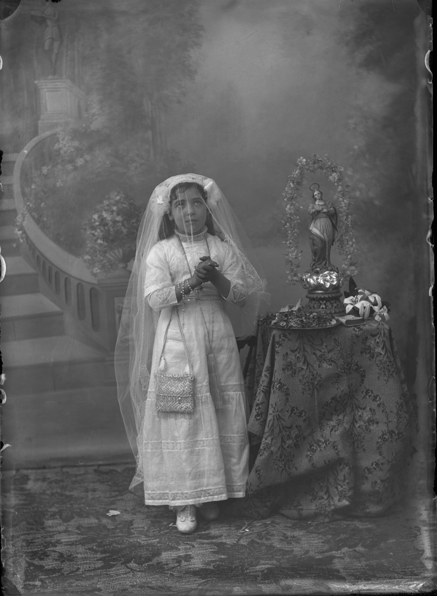 Retrato de estudio de una niña vestida de comunión en actitud de oración junto a una estatuilla de la Virgen