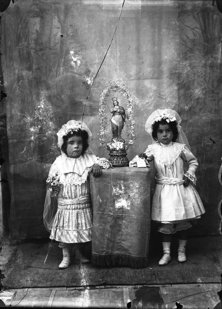 Retrato de estudio de dos niñas con tocado junto a una estatuilla de la Virgen