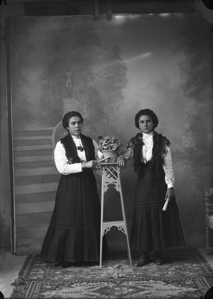 Retrato de estudio de dos mujeres jóvenes con indumentaria similiar junto a una rinconera con maceta
