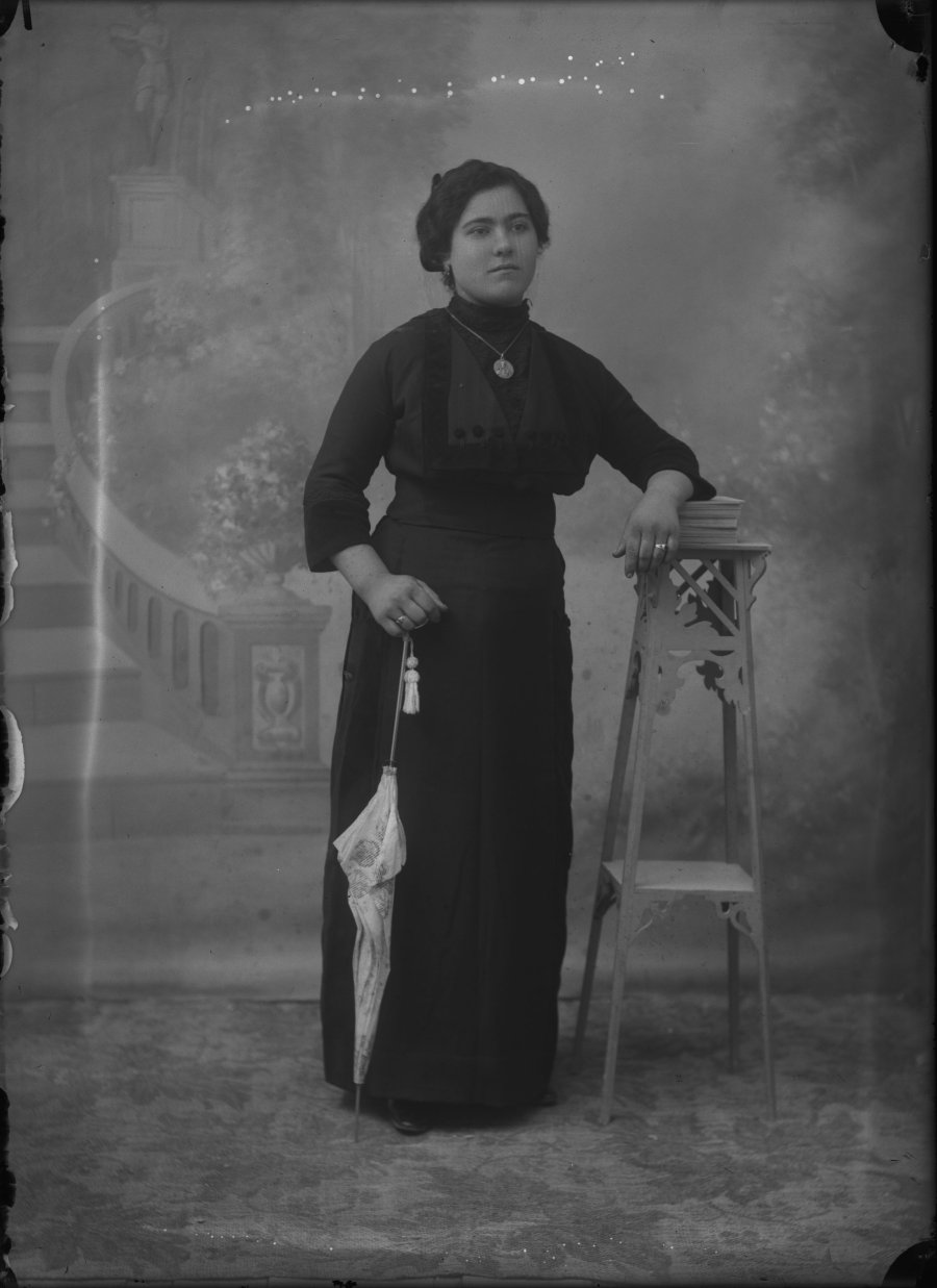 Retrato de estudio de una mujer joven con vestido oscuro, medallón y sombrilla blanca