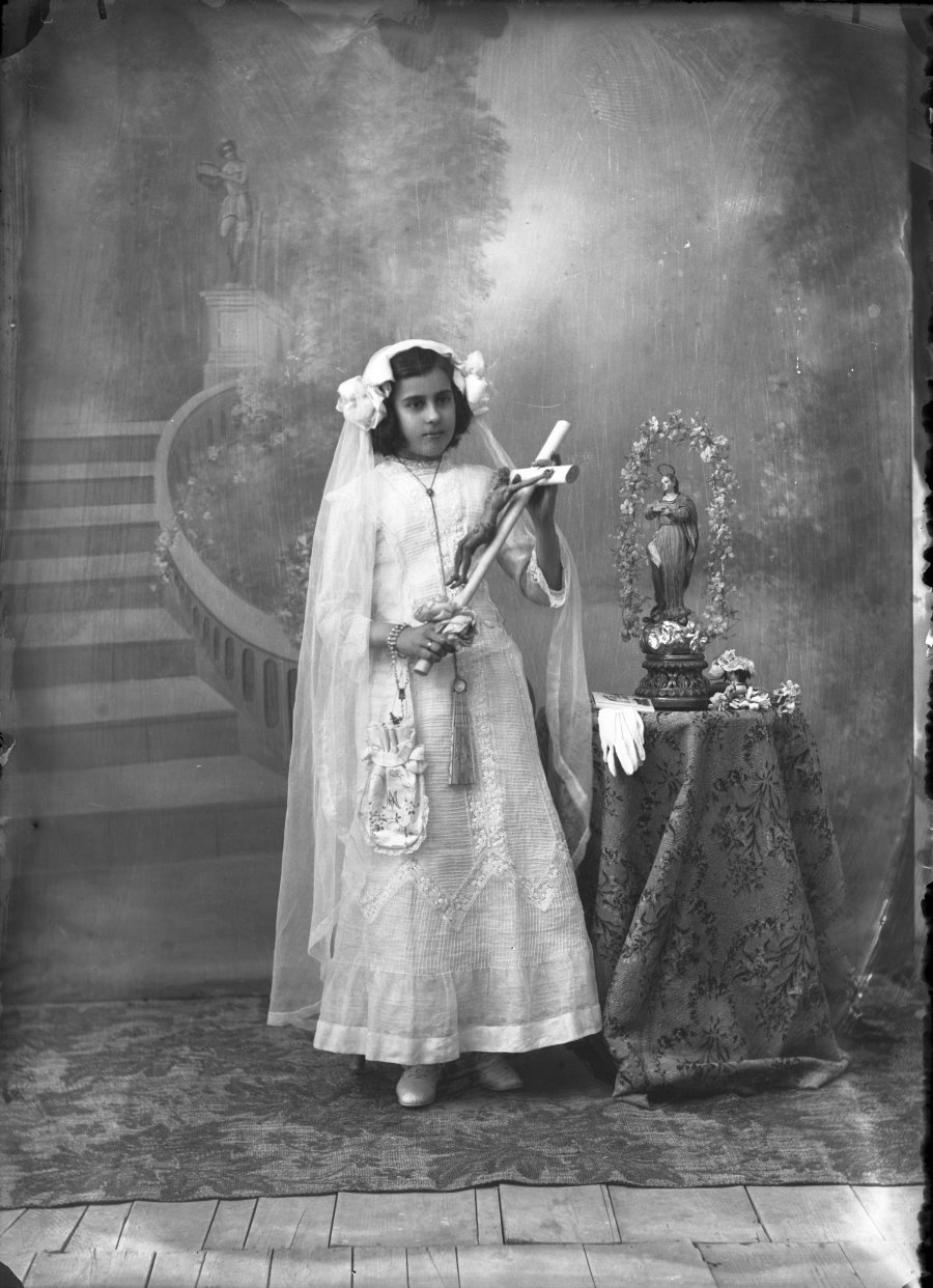 Retrato de estudio de una niña de primera comunión con crucifijo y estatuilla de la Virgen