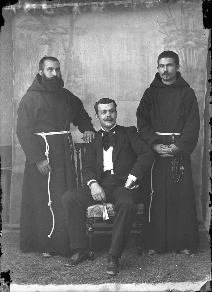 Retrato de estudio de un hombre vestido con traje acompañado por dos frailes capuchinos