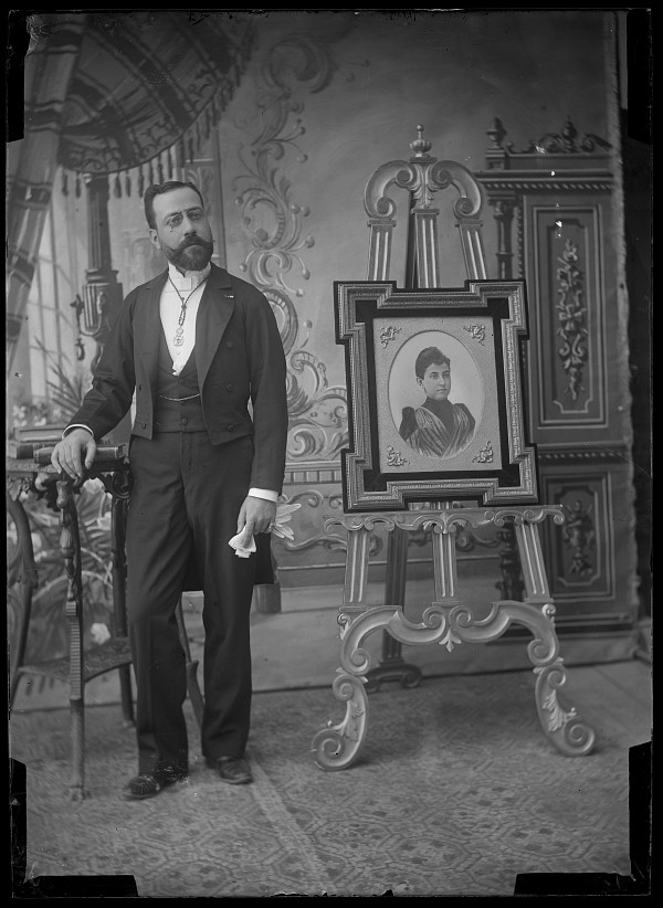 Retrato de estudio de un hombre ataviado con frac junto a un caballete en el figura el retrato de una mujer