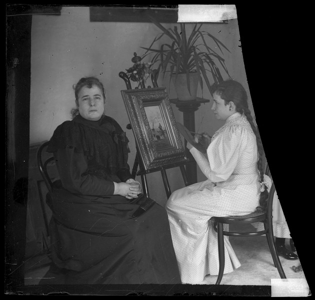 Retrato en interior de una señora sentada junto a muchacha que simula pintar un cuadro
