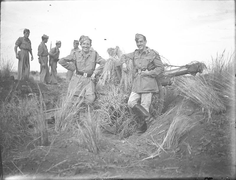 Dos oficiales posando frente a una pieza de artillería camuflada con rastrojos
