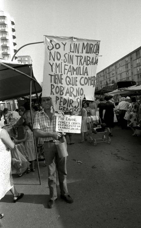 Un parado, con un cartel solicitando ayuda y trabajo, en el tradicional mercado semanal de los jueves de Murcia.