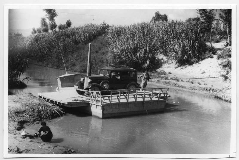 Fotografía de un coche cruzando el río Segura por el Menchú sobre una barcaza de madera.