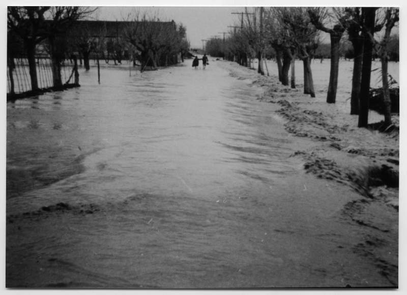 Reproducción de una fotografía del río Segura desbordado con dos personas caminando al fondo.
