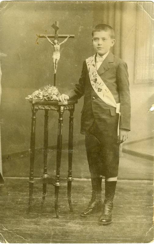 Retrato de niño con traje y banda junto a un crucifijo.