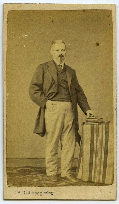 Retrato de estudio de un hombre de pie, con levita, apoyado sobre tres  libros situados sobre una mesa vestida.