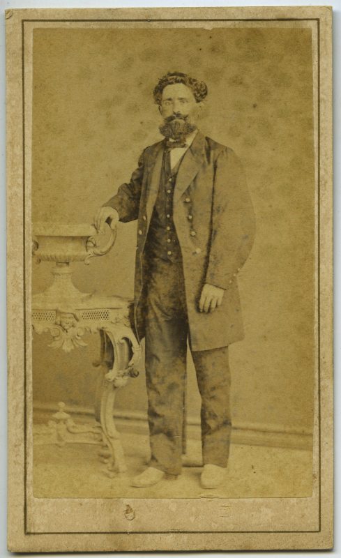Retrato de estudio de un hombre de pie, con levita de botonadura metálica, con la mano derecha apoyada sobre un jarrón de sobremesa.