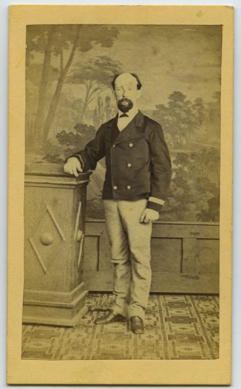 Retrato de estudio de un hombre de pie, con chaqueta corta y galón en la bocamenga, apoyado en un pedestal.