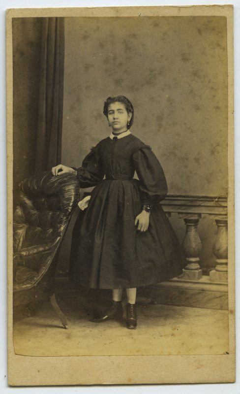 Retrato de estudio de una niña de pie, con vestido negro, apoyada en una butaca.