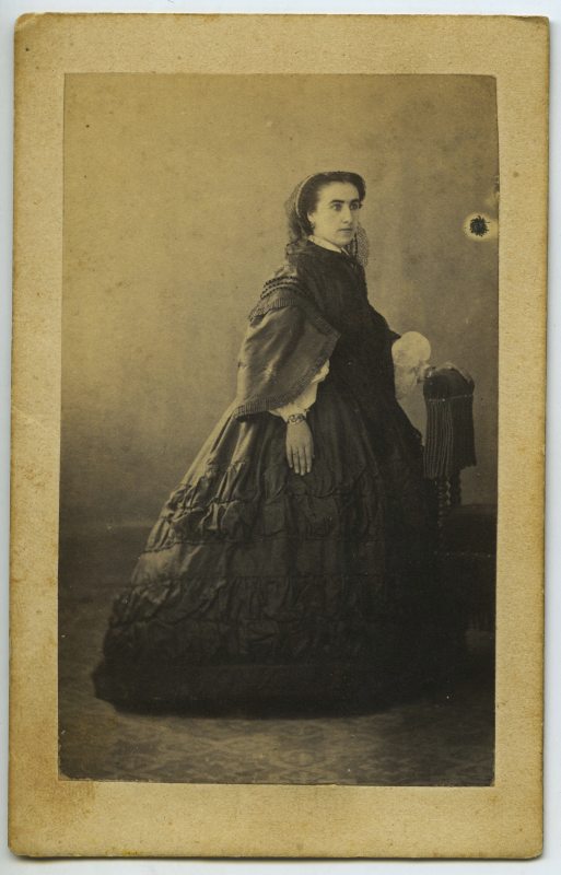 Retrato de estudio de una mujer joven de pie, con vestido negro, apoyada en un sillón.