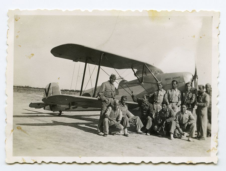 Un grupo de militares y mecánicos posa junto a un biplano en la pista de un aeródromo.